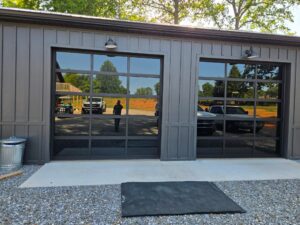 Two glass garage doors on a metal building reflect parked vehicles, trees, and a person standing outside on a gravel surface.
