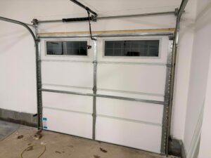 Closed white garage door with rectangular windows at the top, viewed from inside a garage with unfinished flooring and visible garage door opener hardware.