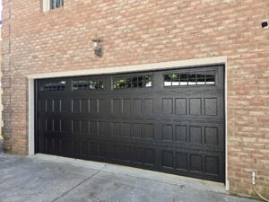 A black garage door with four upper windows is set in a brick wall; a light fixture is mounted above the door.