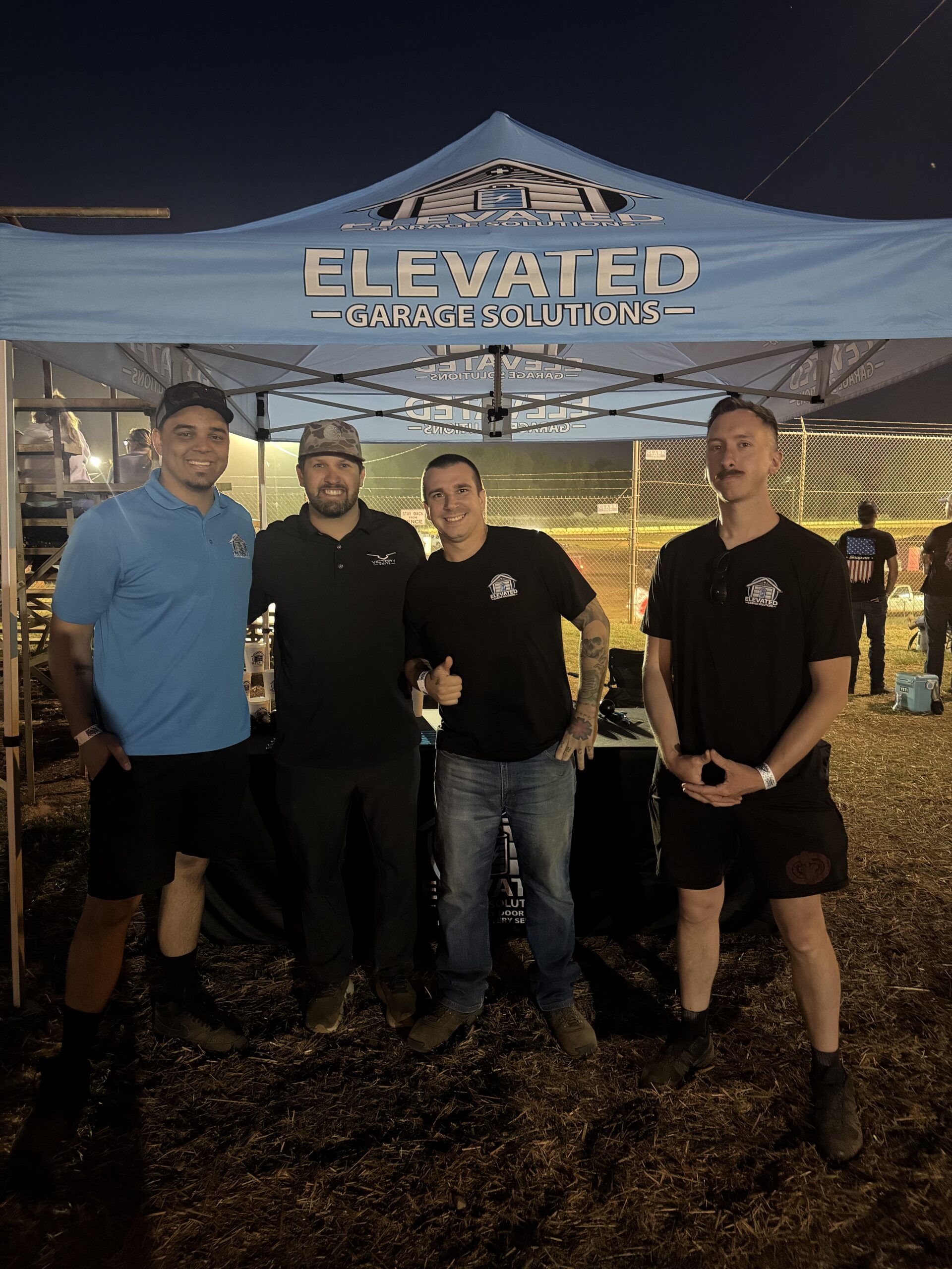 Four men stand under a blue “Elevated Garage Solutions” tent at an outdoor event during the evening.