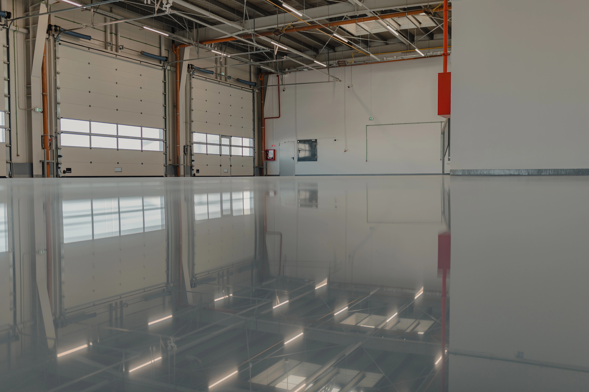 Interior of an empty industrial warehouse with high ceilings, large windows, roller doors, and a polished reflective floor.
