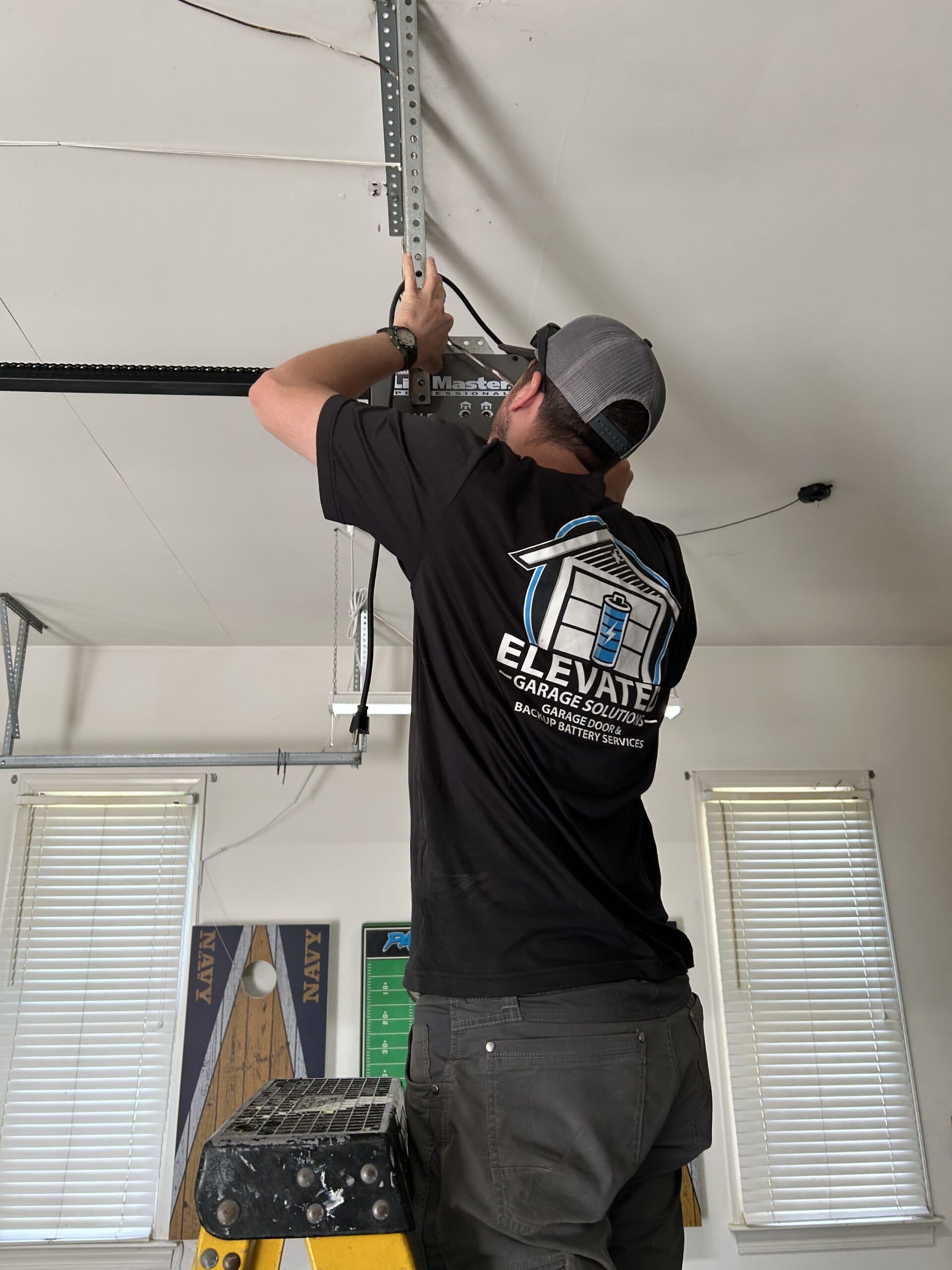 Person in a black “Elevate Garage Solutions” shirt repairs a garage door opener mechanism while standing on a ladder in a well-lit garage.