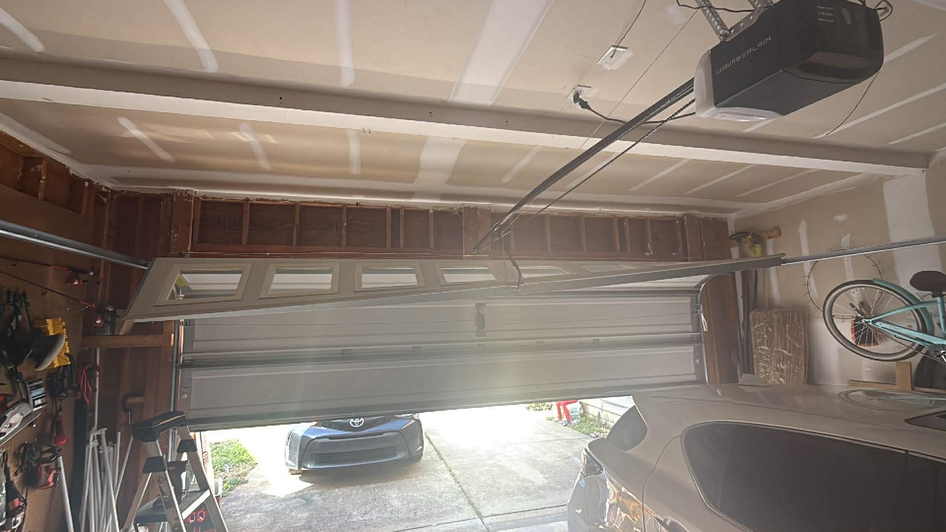 A garage door is partially detached and crooked, with two cars visible—one inside the garage and one parked outside. Tools and a bicycle are mounted on the garage walls.