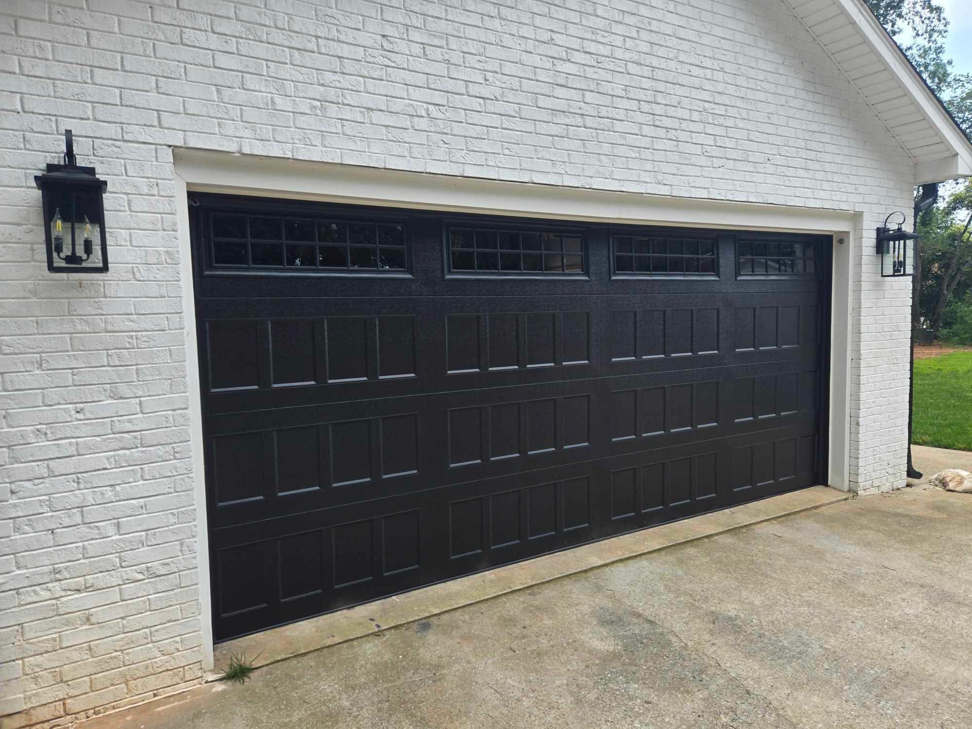 Black paneled garage door with rectangular windows at the top, set in a white brick wall with an exterior lantern-style light fixture on the left side.