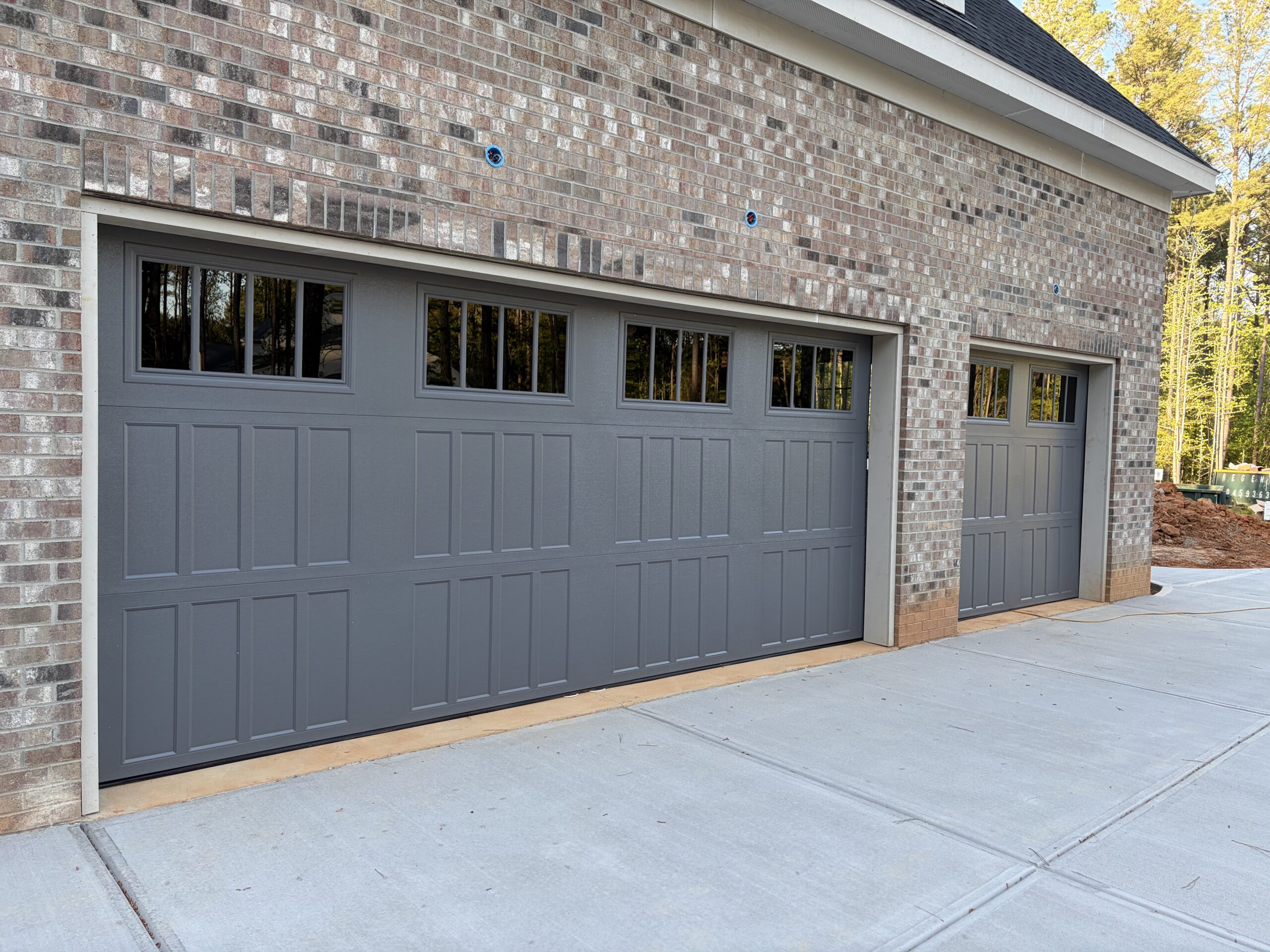 Two gray garage doors with rectangular windows along the top are set into a brick building next to a clean, wide concrete driveway.