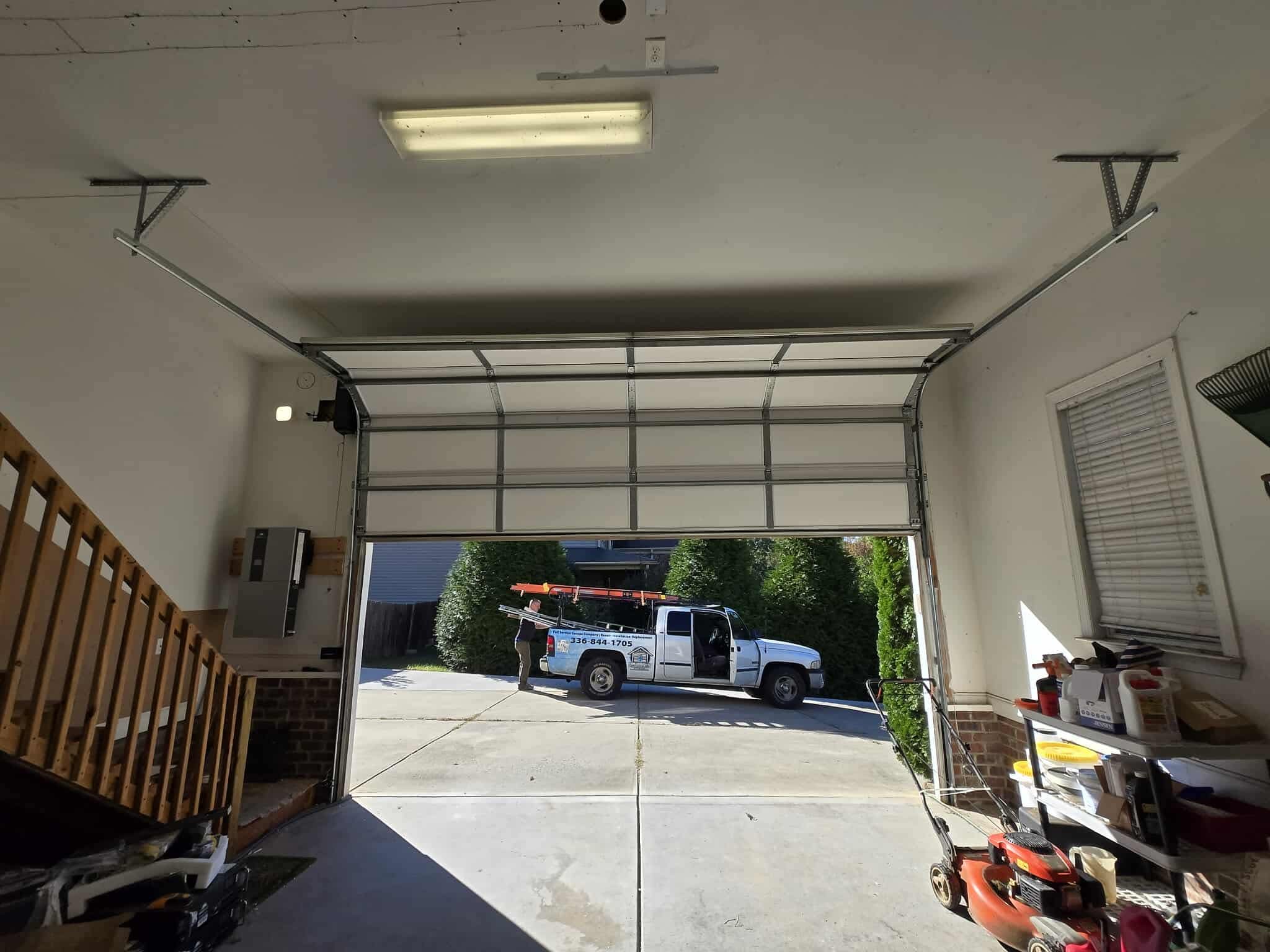 Interior view of an open garage with stairs to the left, lawn equipment on the right, and a pickup truck parked outside in the driveway.
