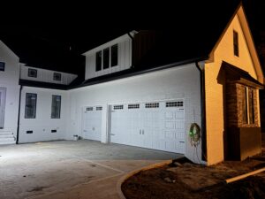 A white two-story house with three garage doors is shown at night, illuminated by exterior lights. A hose is coiled on the right side near the garage.