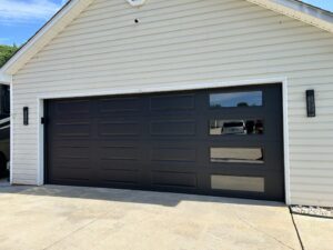 A closed black garage door with rectangular windows on the right side, attached to a beige house with white trim, on a concrete driveway.
