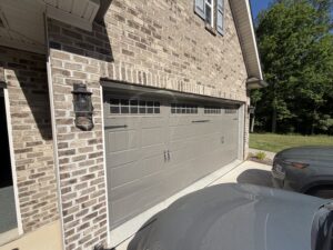 Two closed beige garage doors on a brick house, with two parked gray cars in the driveway. Trees and grass are visible in the background under a clear blue sky.