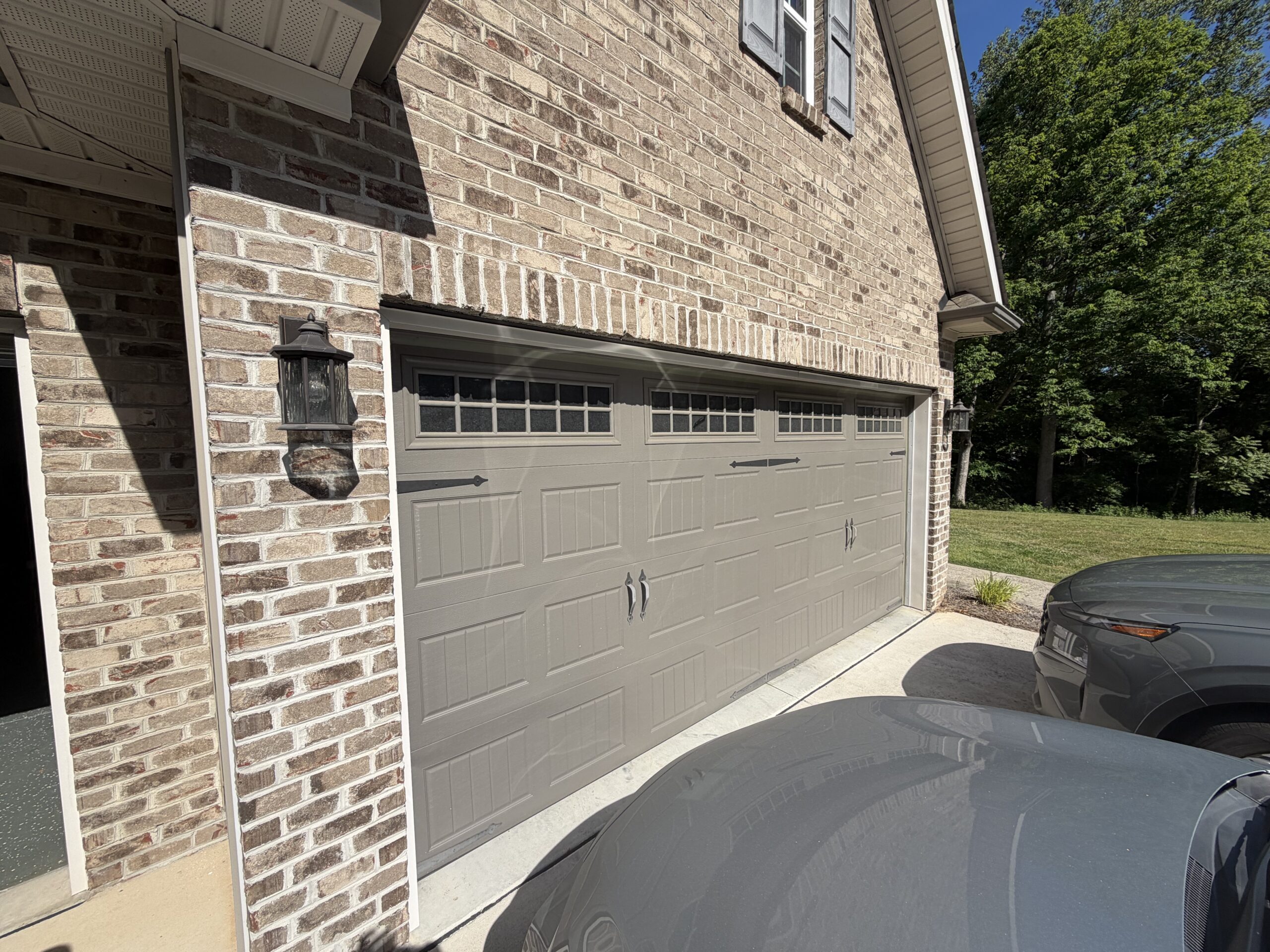 Two closed beige garage doors on a brick house, with two parked gray cars in the driveway. Trees and grass are visible in the background under a clear blue sky.
