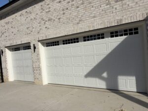 Two closed white garage doors with small windows at the top, set in a light brick wall, with sunlight casting a large shadow across the right side.