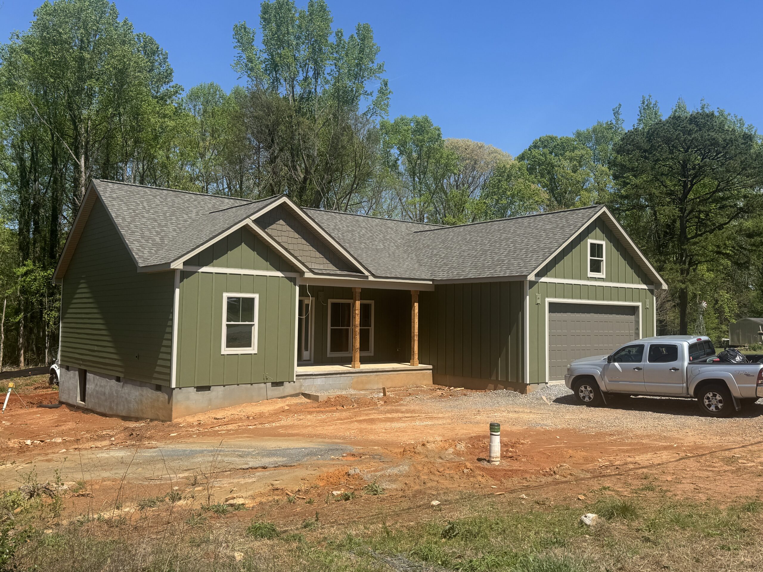 A newly constructed green house with a front porch and attached garage, surrounded by red dirt and trees, with two parked trucks in the driveway.