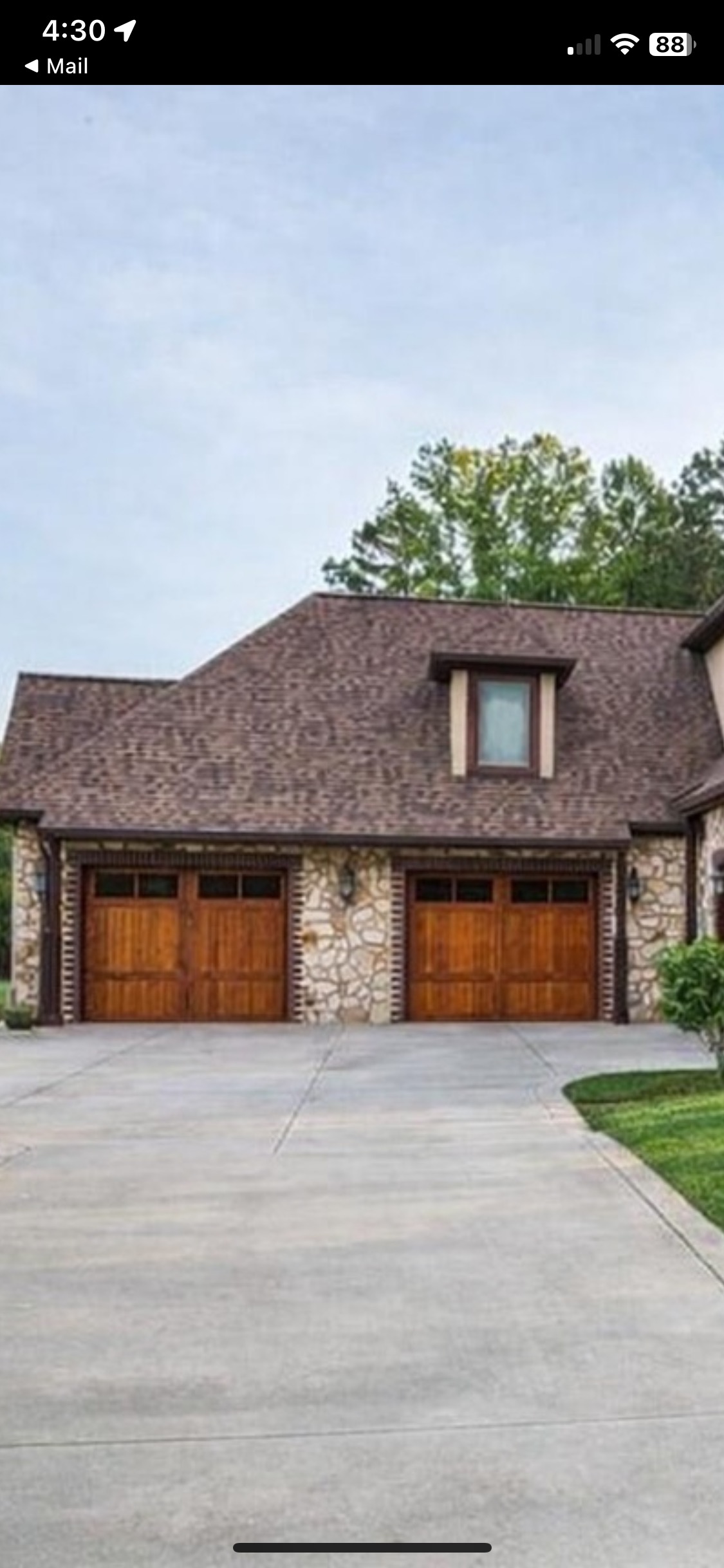 A two-car garage with wooden doors is attached to a stone and brick house with a brown shingle roof and a dormer window, set beside a concrete driveway and a small lawn.